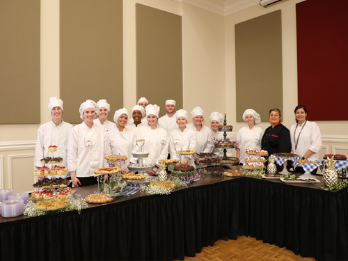 Group of students in chef coats and hats with their instructor behind a table of food