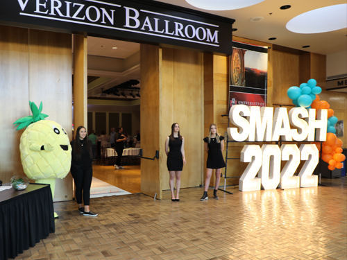 Three students standing outside a ballroom with a person in a pineapple costume and a sign reading SMASH 2022